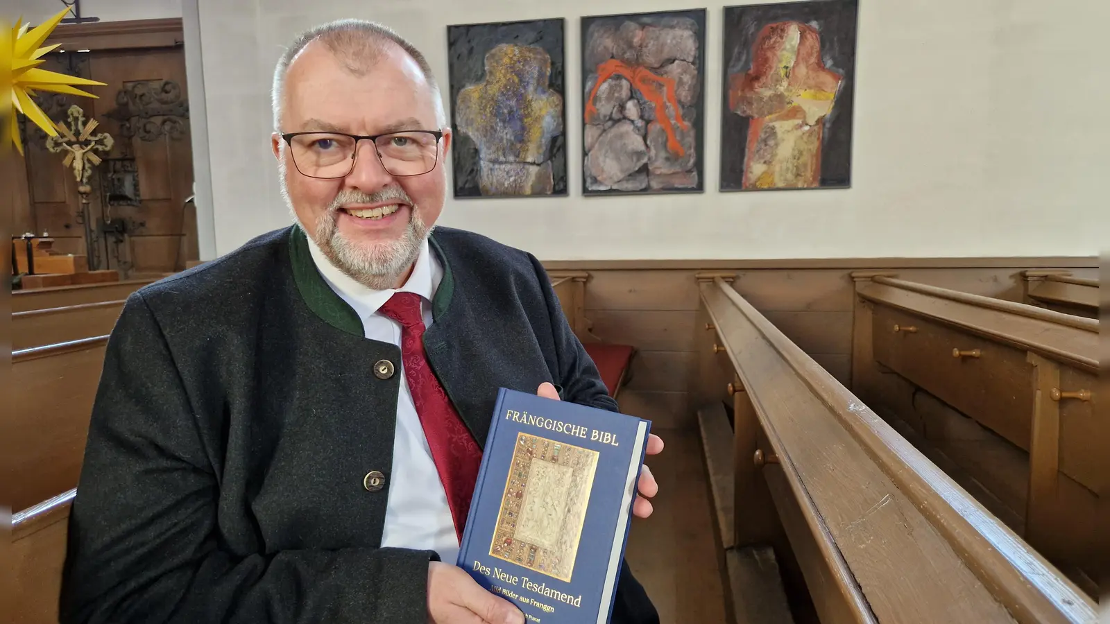 Pfarrer Claus Ebeling zeigt die Neuauflage der Fränggischen Bibl. Motive wie das Triptychon von Waltraut Beck zum Thema „Kain und Abel“ (im Hintergrund) werden für das Alte Testament gesucht. (Foto: Andrea Walke)