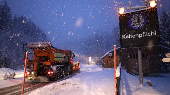 Ein Schild weist auf die Schneekettenpflicht am Riedbergpass im bayerischen Landkreis Oberallgäu hin, während daneben ein Räumfahrzeug vom Winterdienst den Schnee von der Straße räumt. (Symbolbild) (Foto: Davor Knappmeyer/dpa)
