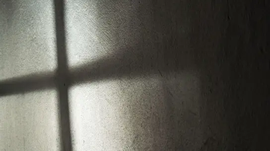 Ein Fenster wirft seinen Schatten auf die Wand. Es entsteht ein dunkles Kreuz. (Foto: Annette Riedl/dpa/Archivbild)