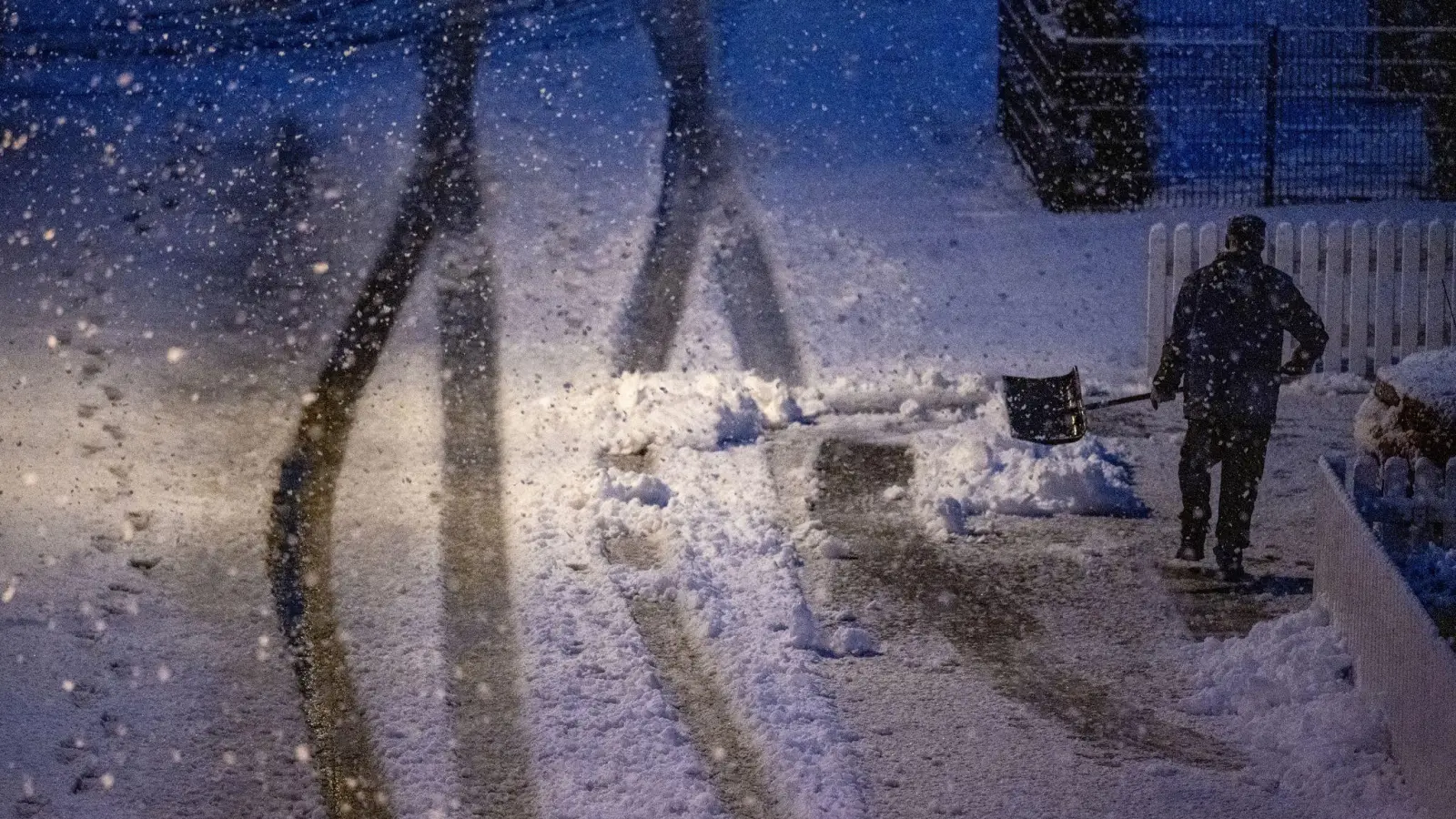 Ein Mann geht am frühen Morgen mit einer Schneeschaufel auf einer zugeschneiten Straße. (Foto: Armin Weigel/dpa)