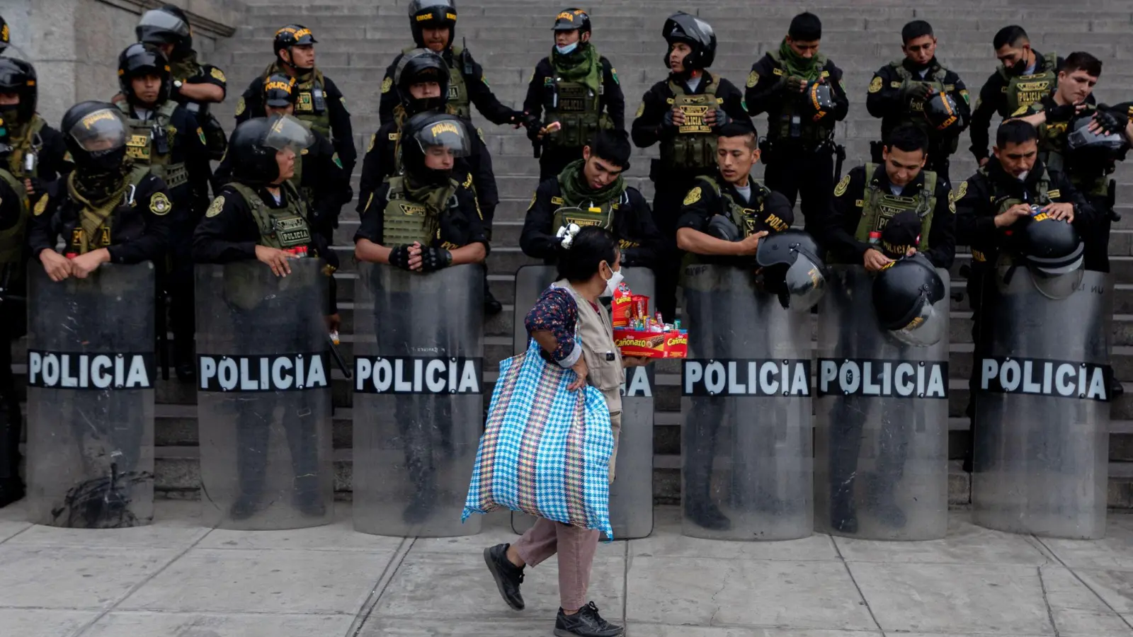 Gegensätze in Perus Hauptstadt Lima: Eine Frau läuft mit Süßigkeiten in der Hand an einer mit Schutzschilden und Helmen ausgestatteten Gruppe von Polizisten vorüber. (Foto: Lucas Aguayo Araos/dpa)