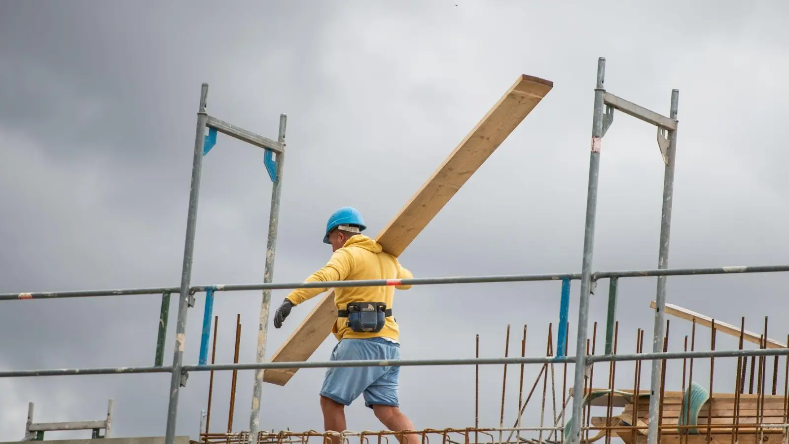 Ein Bauarbeiter trägt auf einer Baustelle auf einem Hausdach ein Brett. (Foto: Christophe Gateau/dpa)