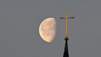 Der Mond im Morgenlicht hinter einem Kreuz auf einem Kirchturm. (Foto: Bernd Weißbrod/dpa)