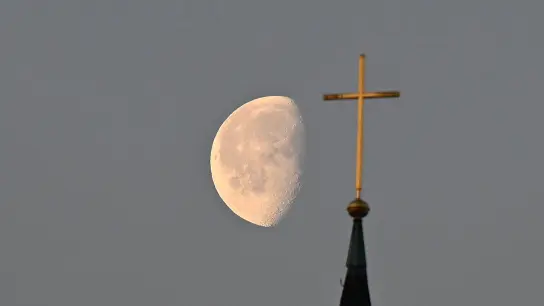 Der Mond steht im Morgenlicht hinter einem Kreuz auf einem Kirchturm. (Foto: Bernd Weißbrod/dpa/Symbolbild)