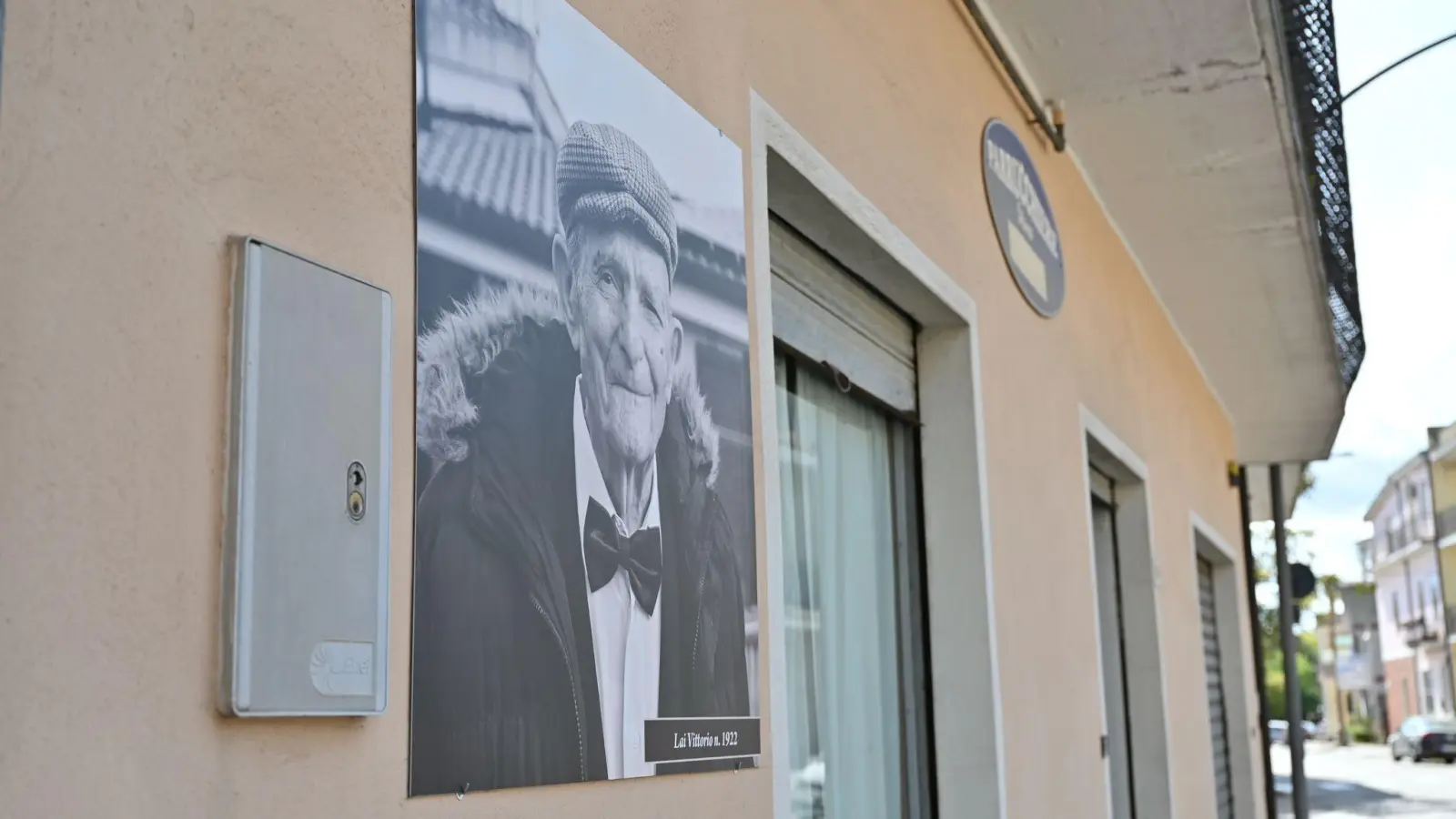 An der Fassade der Häuserin Perdasdefogu hängen Schwarz-Weiß-Fotos der Hundertjährigen aus dem Dorf mit deren Namen und Geburtsjahr. Auf diesem Bild ist Vittorio Lai zu sehen. (Foto: Johannes Neudecker/dpa)