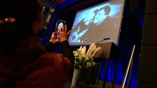 Eine Frau fotografiert mit ihrem Smartphone die Bühne mit Blumenstrauß und einem Foto des Verstorbenen auf einer Leinwand. (Foto: Jonas Walzberg/dpa)