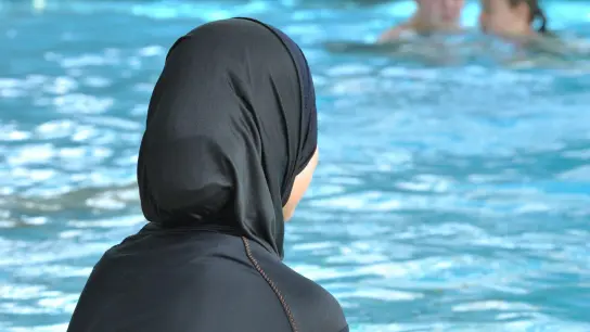 Eine muslimische Schülerin sitzt in einem Ganzkörperbadeanzug (Burkini) am Rande eines Schwimmbeckens. In Frankreich ist erneut ein Streit um Burkinis ausgebrochen. (Foto: Rolf Haid/dpa)