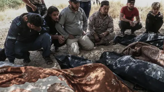Trauernde sitzen 2021 neben eingehüllten Leichen unter Bäumen in Idlib und warten darauf, dass Kampfflugzeuge abziehen. (Foto: Anas Alkharboutli/dpa)
