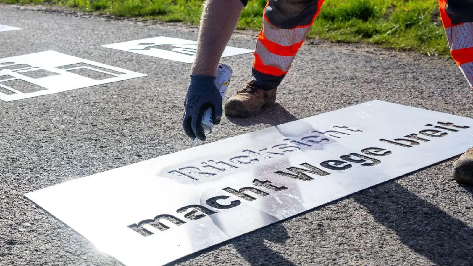 Der Mitarbeiter eines Bauhofs sprüht den Schriftzug „Rücksicht macht Wege breit“ auf einen Feldweg, während eine Radfahrerin auf ihn zufährt. (Foto: Pia Bayer/dpa)
