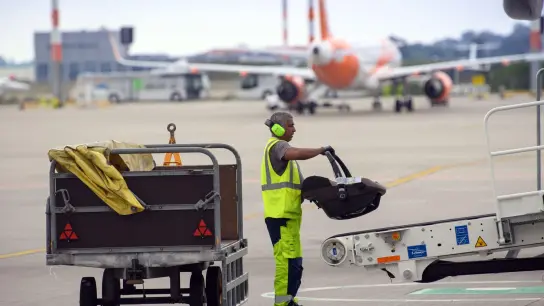 Ein Mitarbeiter der WISAG Aviation lädt auf dem Vorfeld des Flughafen BER „Willy Brandt“ einen Kindersitz auf das Gepäck-Transportband am Flugzeug. (Foto: Soeren Stache/dpa-Zentralbild/dpa)