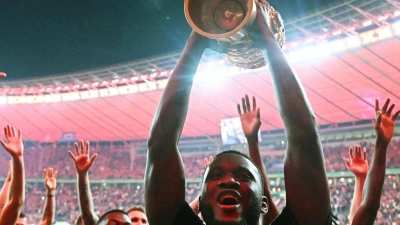 Leverkusens Victor Boniface (r) hält den Pokal jubelnd hoch, während seine Teamkollegen feiern. (Foto: Federico Gambarini/dpa)