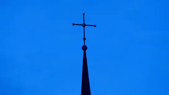 Das Kreuz einer Kirche ist in der Abenddämmerung zu sehen. (Foto: Friso Gentsch/dpa/Symbolbild)