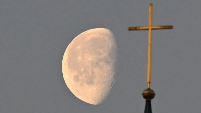 Der Mond steht im Morgenlicht hinter einem Kreuz auf einem Kirchturm. (Foto: Bernd Weißbrod/dpa/Symbolbild)