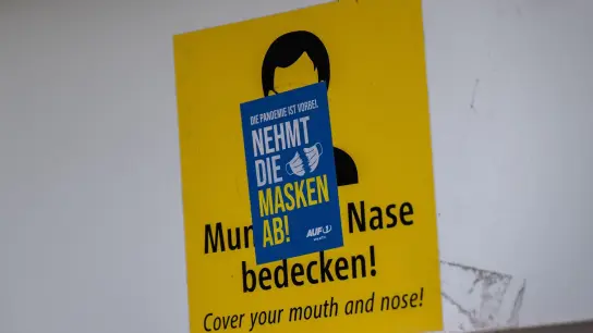 Ein Aufkleber mit der Aufschrift „Nehmt die Masken ab!“ klebt über einem Schild, das zum Tragen einer Mund-Nasen-Maske auffordert. Die Mehrheit der Deutschen ist einer neuen Umfrage zufolge weiter für eine Isolationspflicht für Corona-Infizierte. (Foto: Peter Kneffel/dpa)