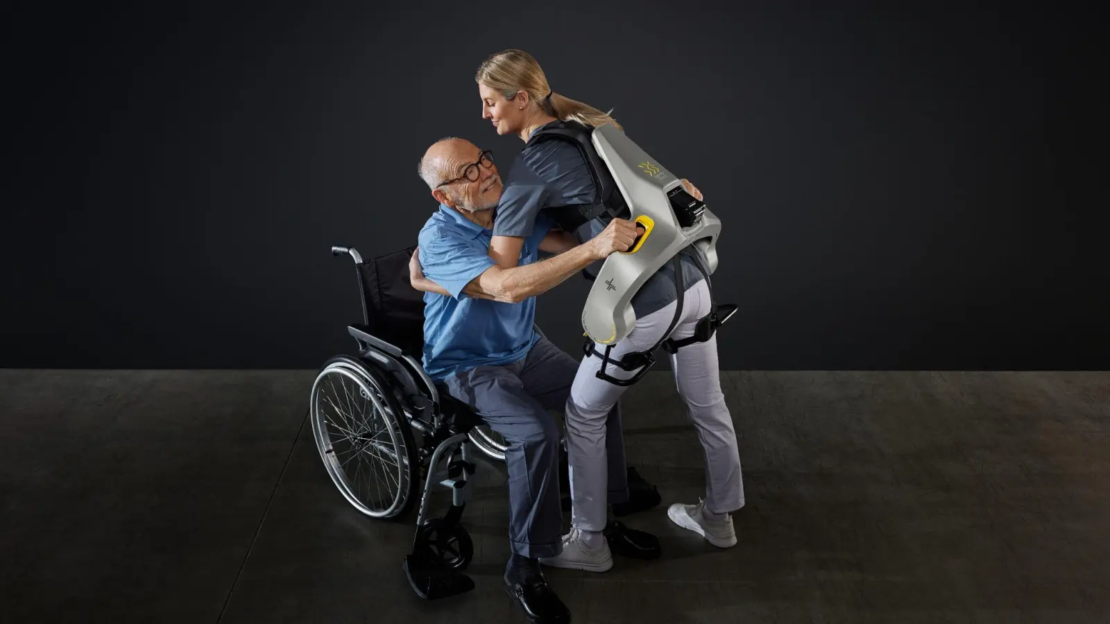 Eine Frau hebt mit Hilfe eines intelligenten Kraftanzugs der Firma German Bionic einen Mann aus einem Rollstuhl. (Foto: German Bionic/dpa)
