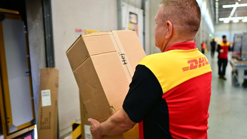 Ein Paketbote schleppt in einem DHL-Standort ein schweres Paket. (Archivbild) (Foto: Sebastian Christoph Gollnow/dpa)