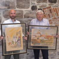 Die Stiftsbasilika und der Storchenturm in Aquarell: Diese beiden Bilder, die Herriedens Bürgermeister Patrick Peters (links) und Altbürgermeister Werner Herzog präsentieren, gehören zur Sammlung, die die Stadt nun ihr Eigen nennen darf. (Foto: Jasmin Kiendl)