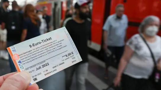 Ein 9-Euro-Ticket für Juli 2022 wird am Hauptbahnhof hochgehalten, während Zugreisende aus einer Regionalbahn aussteigen. (Foto: Arne Dedert/dpa)