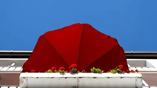 Ein roter Sonnenschirm und rote Geranien sind auf einem Balkon zu sehen. (Foto: Sven Hoppe/dpa)