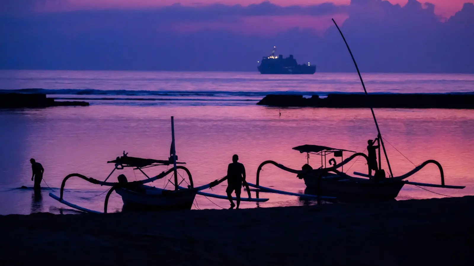 Männer befestigen vor Sonnenaufgang am Tag vor Beginn des G20-Gipfels am Strand von Nusa Dua Boote, wobei im Hintergrund ein Schiff der indonesischen Marine zu sehen ist. (Foto: Christoph Soeder/dpa)