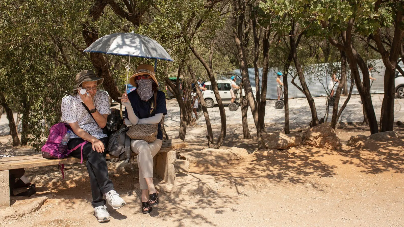 Touristen sitzen unter Bäumen am Eingang der Akropolis in Athen. (Foto: Socrates Baltagiannis/dpa)