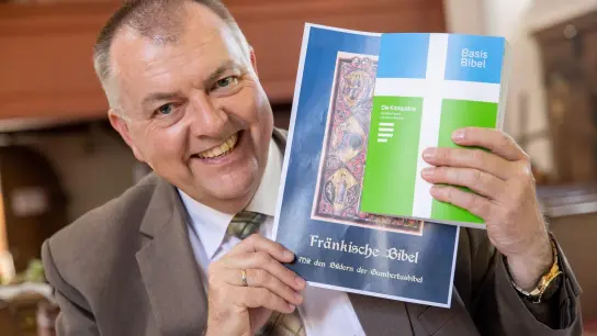 Pfarrer Claus Ebeling hält einen Entwurf für den Buchdeckel einer Mundart-Bibel neben einer herkömmlichen Bibel. (Foto: Daniel Karmann/dpa/Archivbild)