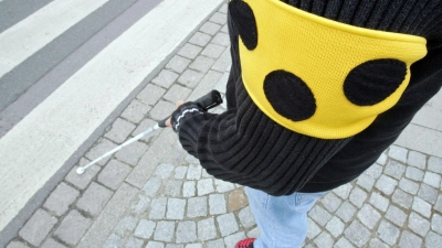 Ein blinder Mann geht mit seinem weißen Taststock und einer Armbinde mit drei Punkten über eine Straße in Hannover. (Foto: Patrick Lux/dpa)