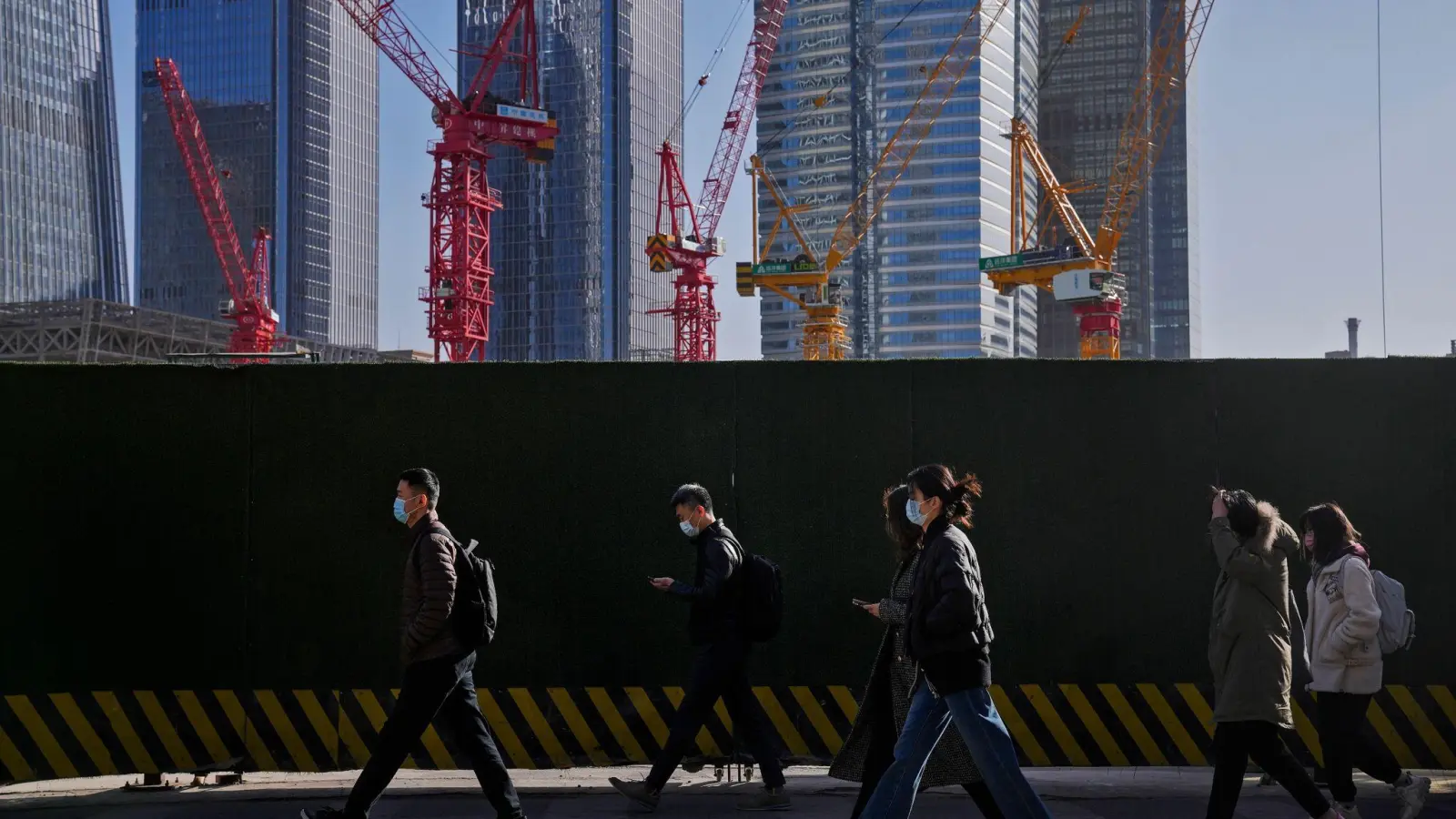 Menschen mit Masken gehen an Baukränen in Pekings Geschäftsviertel vorbei. Erste Wirtschaftszahlen nach der Abkehr Chinas von seiner strikten Corona-Politik zeigen eine moderate Erholung der Konjunktur. (Foto: Andy Wong/AP)