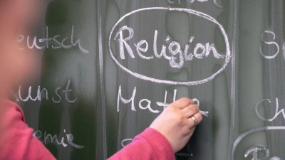 Eine Lehrerin schreibt mit Kreide einen Stundenplan mit Religionsunterricht an eine Tafel. (Foto: Friso Gentsch/dpa)