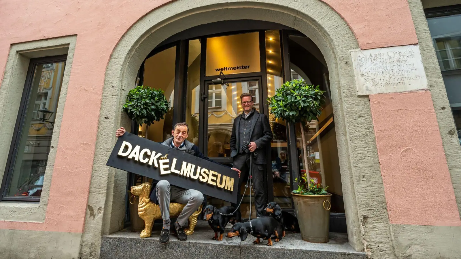 Seppi Küblbeck (l) und Oliver Storz stehen mit ihren drei Dackeln vor dem zukünftigen Dackelmuseum. (Foto: Armin Weigel/dpa)