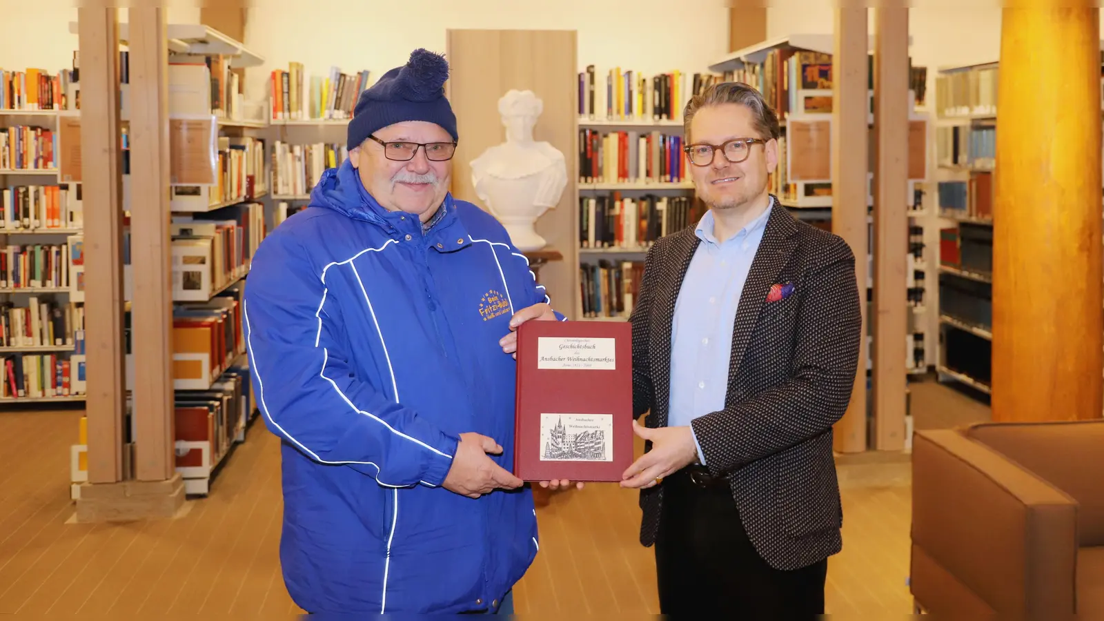 Fritz Heubeck (links) übergibt Bibliotheksleiter Christian Mantsch ein Exemplar seines „Chronologischen Geschichtsbuchs des Ansbacher Weihnachtsmarktes”. (Foto: Oliver Herbst)