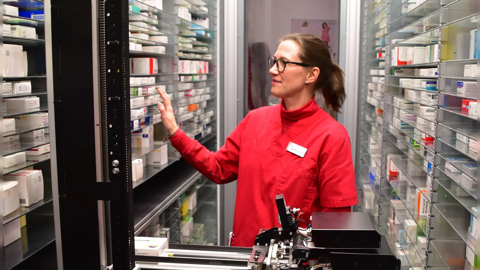 Annette Pauli erklärt, wie Arzneimittel per Zufallsprinzip automatisch auf freie Fächer verteilt werden. Die Packungen werden von dem Schieber zunächst horizontal zu den passenden Etagen gebracht und dann nach oben oder unten vor das per Zufall ausgewählte freie Fach bugsiert. (Foto: Kurt Güner)