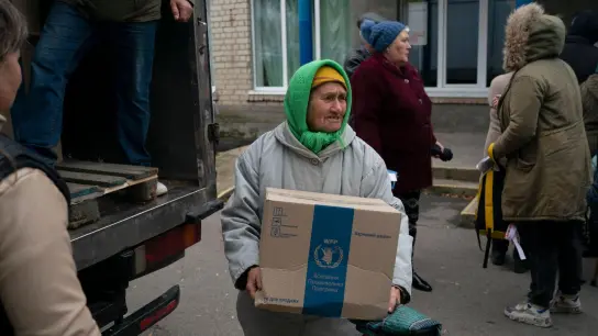 Eine ältere Frau trägt eine Kiste, während in dem Dorf Mychajlo-Lukaschewe humanitäre Hilfsgüter verteilt werden. (Foto: Leo Correa/AP/dpa)