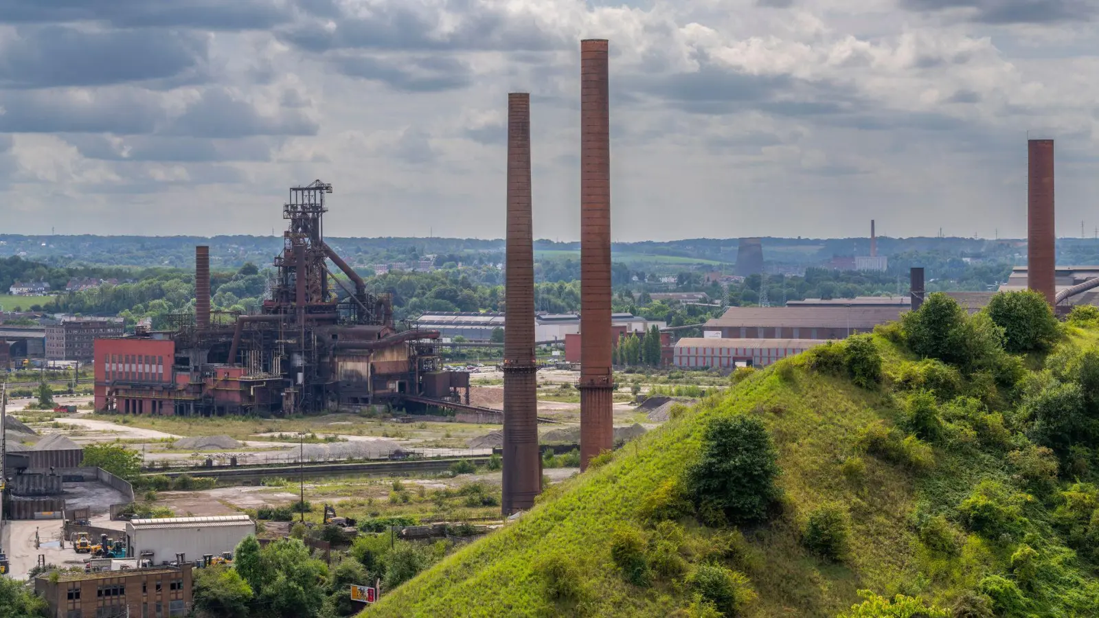 Industriegebiet von Charleroi: Blick über eine bewachsene Halde des Kohleabbaus am Wanderweg „Boucle Noire“, hinten Schornsteine und ein ehemaliger Hochofen eines Stahlwerks. (Foto: Oliver Gerhard/dpa-tmn)