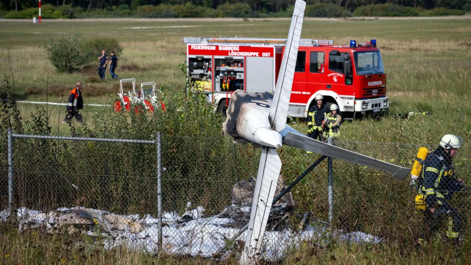 Trümmerteile eines Flugzeugs hängen in einem Zaun, während Einsatzkräfte der Feuerwehr an der Absturzstelle stehen. (Foto: Pia Bayer/dpa)