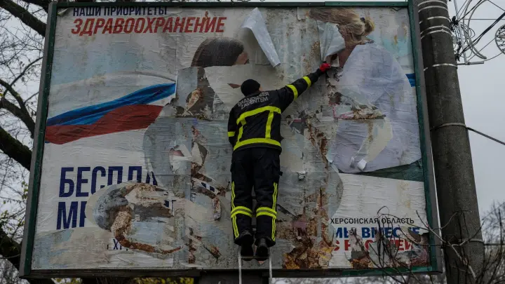 Ein ukrainischer Feuerwehrmann reißt in der kürzlich befreiten Stadt Cherson eine russisches Werbeplakat von einer Werbetafel. (Foto: Bernat Armangue/AP/dpa)