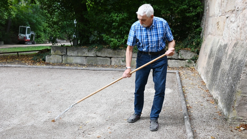 Karl Springer pflegt den Boule-Platz im Stadtgraben in Ansbach. Er muss aktuell immer wieder Löcher und Furchen mithilfe eines Rechens auffüllen. (Foto: Luca Paul)