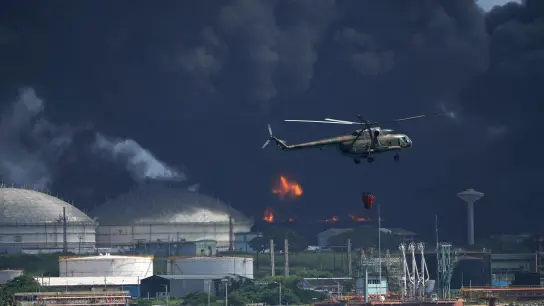 Ein mit Wasser beladener Hubschrauber fliegt über die Supertanker-Basis Matanzas, von der dunkler Rauch und Flammen aufsteigen, während Feuerwehrleute versuchen, den Brand zu löschen. (Foto: Ramon Espinosa/AP/dpa)