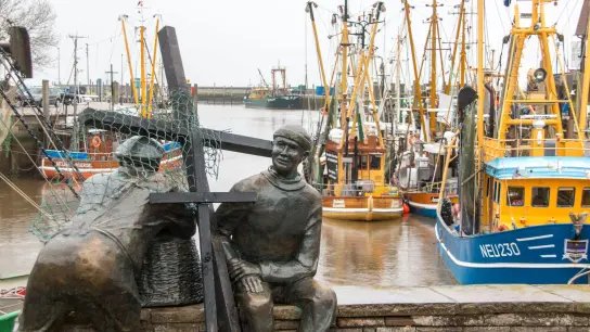 Schwarze Holzkreuze lehnen an der Bronezeskulptur „Alt- und Jungfischer“ im Hafen in Neuharlingersiel. Das Fischen mit Grundschleppnetzen steht in der Kritik. (Foto: Lennart Stock/dpa)