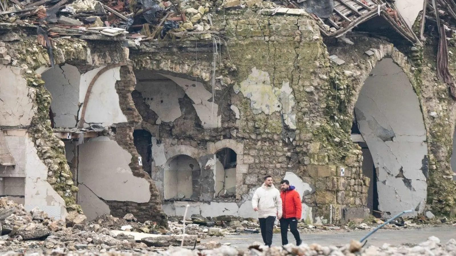 Zwei Männer gehen an einer völlig zerstörten Häuserzeile in Antakya vorbei. Im Zentrum der Stadt waren bei dem Beben vor einem Jahr unzählige Häuser zerstört oder beschädigt worden. (Foto: Boris Roessler/dpa)