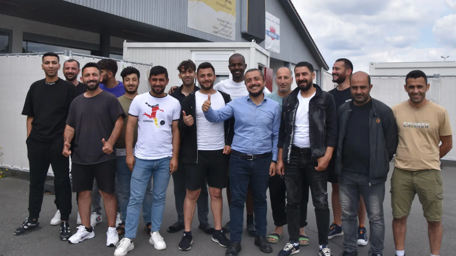 Daumen hoch fürs Team4Scheinfeld zeigt Onur Derin (vorne, im blauen Hemd). Das Foto vor dem Eingang zur Notunterkunft zeigt knapp die Hälfte der Mitstreiter, die sich mit verschiedenen Tätigkeiten ins Stadtleben einbringen wollen. (Foto: Andreas Reum)