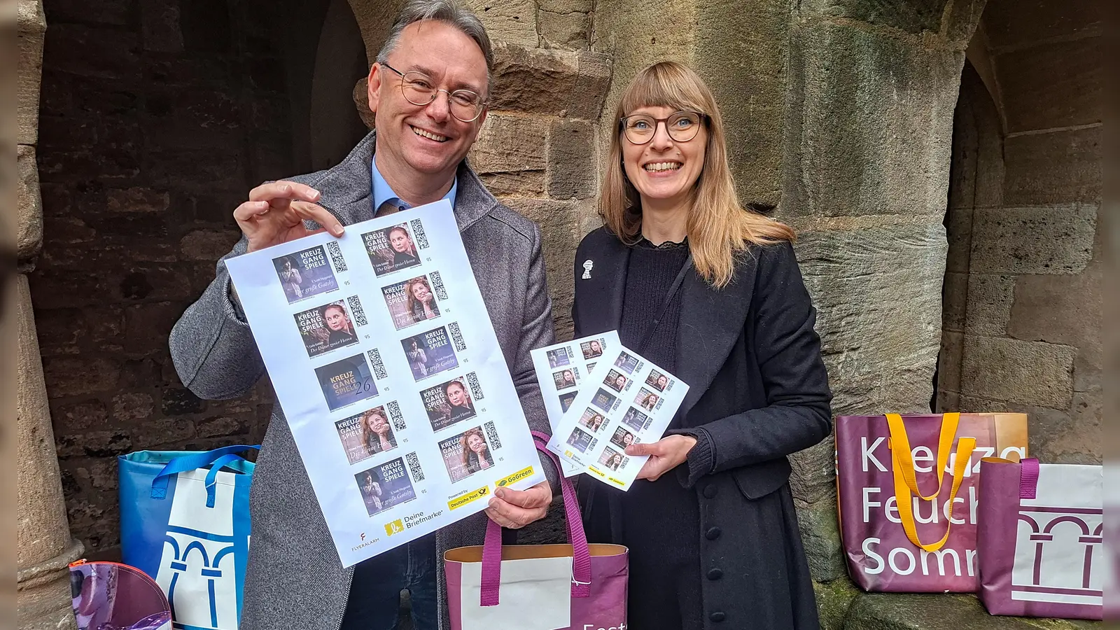 Ab sofort im Kulturbüro erhältlich: limitierte Sonderbriefmarken mit exklusiven Motiven der Kreuzgangspiele 2026 und die neue Kollektion der Kreuzgangspiele-Taschen. Herbert Lindörfer, Vorsitzender des Kreuzgangspiele-Fördervereins, zeigt die Marken in vergrößerter Darstellung, Kulturbüro-Leiterin Dr. Maria Wüstenhagen präsentiert den Zehner-Bogen im Originalformat. (Foto: Stadt Feuchtwangen/Rebecca Weber)